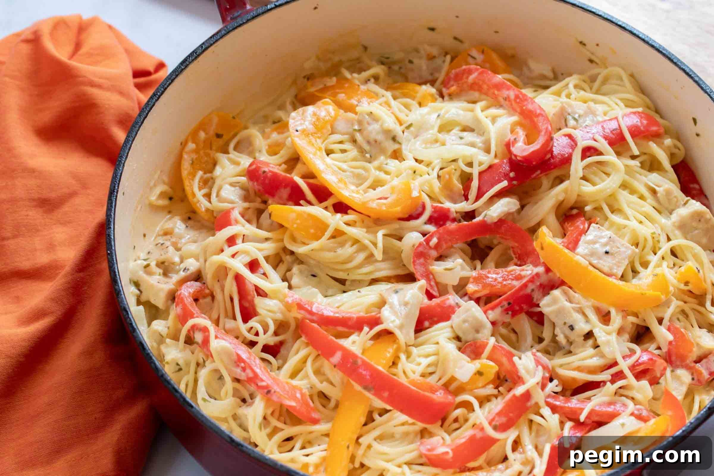 Pasta with chicken, sliced onion, and strips of red and yellow bell peppers cooked in a white sauce in a skillet.
