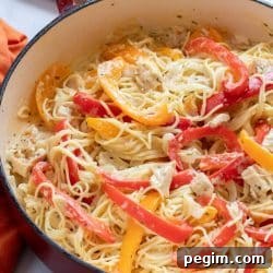 Pasta with chicken, sliced onion, and strips of red and yellow bell peppers cooked in a white sauce in a skillet.