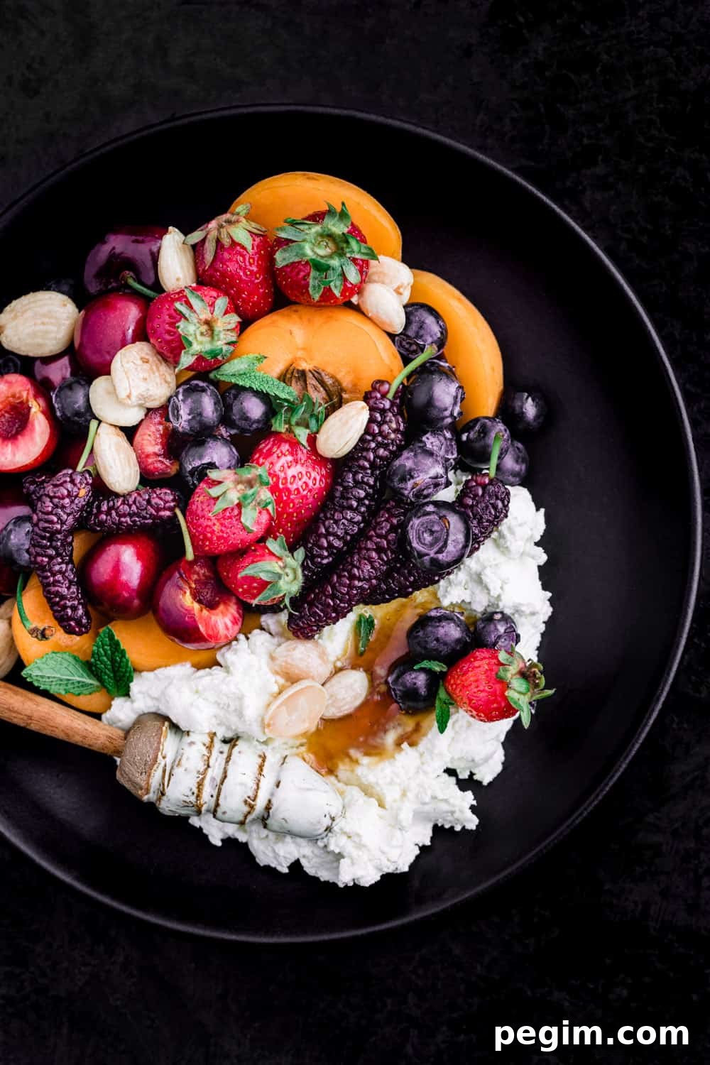 Summer fruit with fresh cheese, almonds and honey on a black plate; overhead shot on a black background.