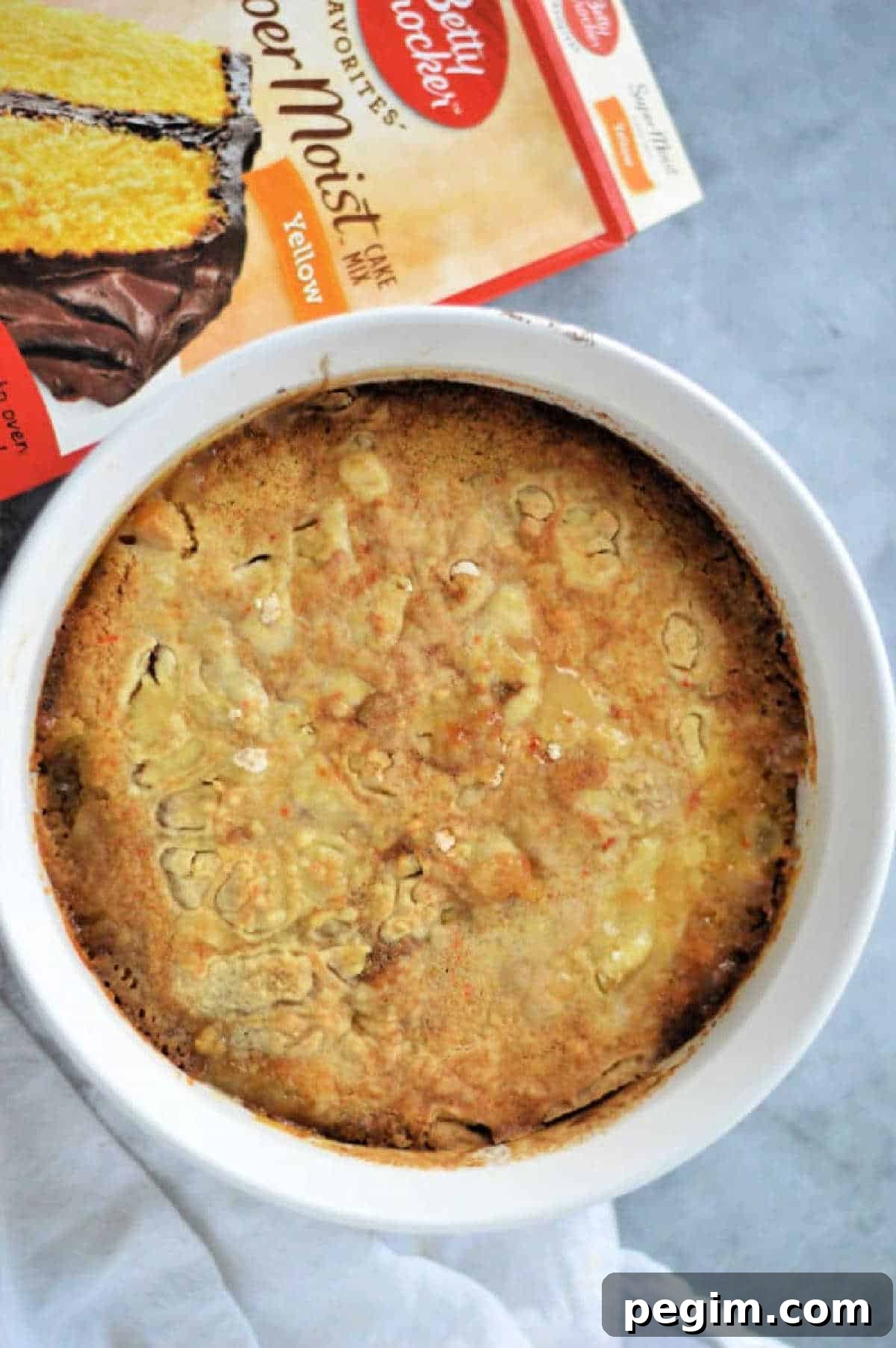 Old-Fashioned Peach Cobbler Made with Cake Mix 4 A peach cobbler with cake mix is baked in a white dish, sitting next to a Betty Crocker yellow cake mix box on a gray surface.