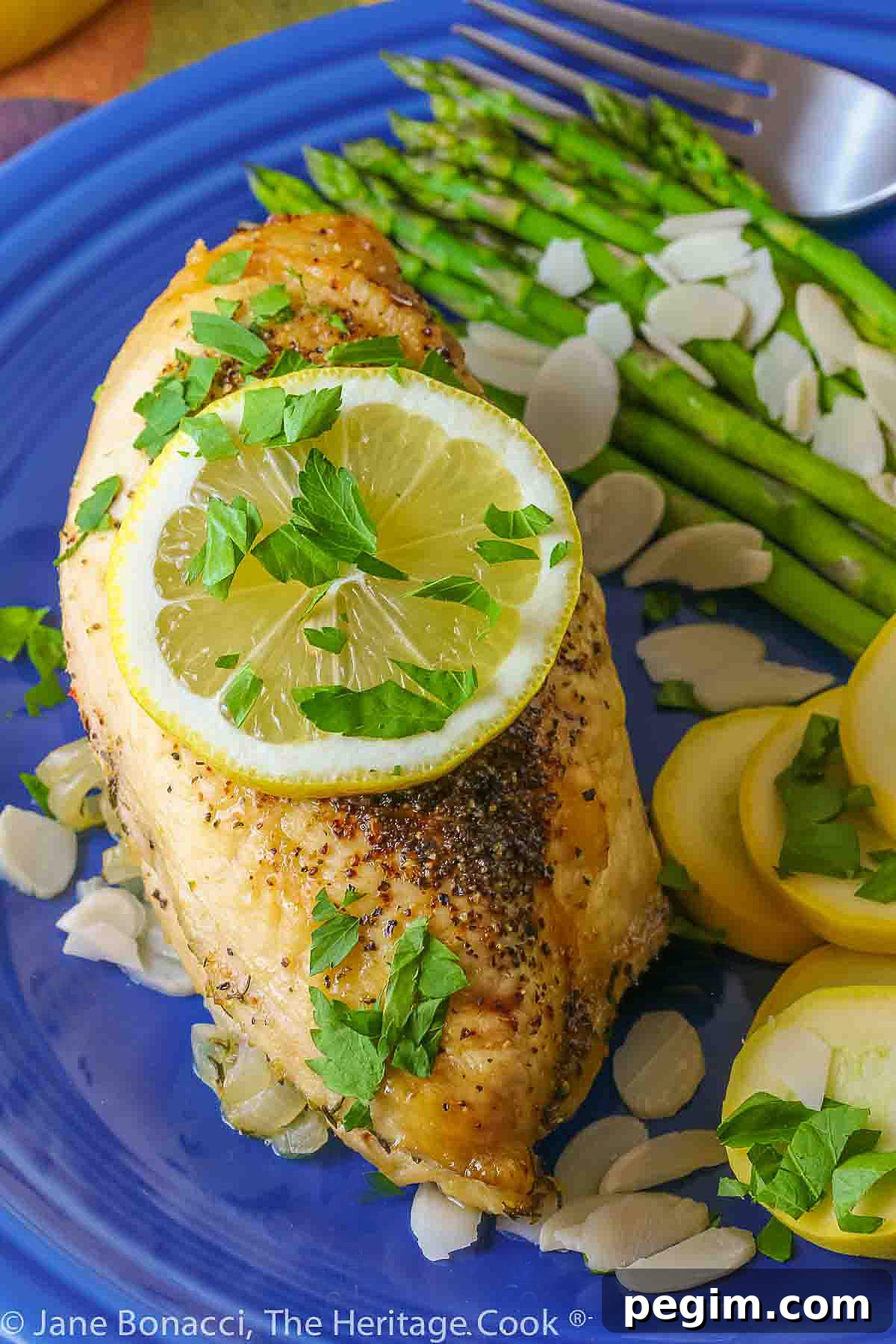 Large baked lemon chicken breast with a slice of lemon on top and green asparagus next to yellow squash on a blue plate © 2024 Jane Bonacci, The Heritage Cook. 