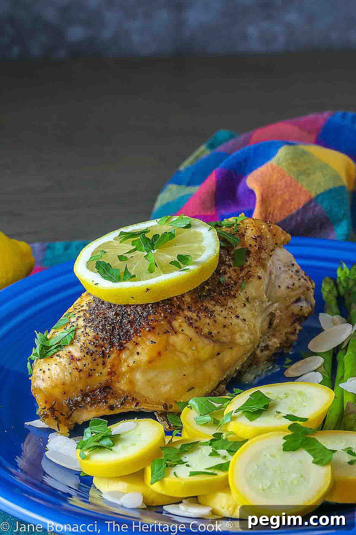 Large baked lemon chicken breast with a slice of lemon on top and green asparagus next to yellow squash on a blue plate © 2024 Jane Bonacci, The Heritage Cook. 