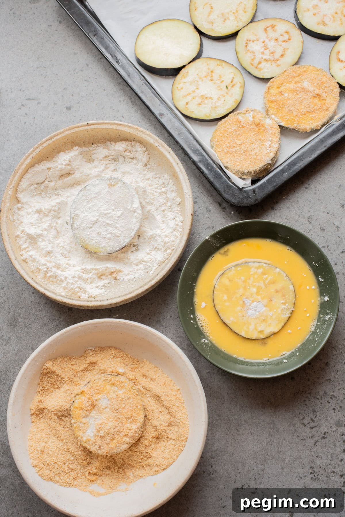 Crispy Baked Eggplant Parmesan Recipe 5 An overhead image of three bowls: one with egg mixture, one with breadcrumbs, and one with flour. With an eggplant slice drenched in each of them.