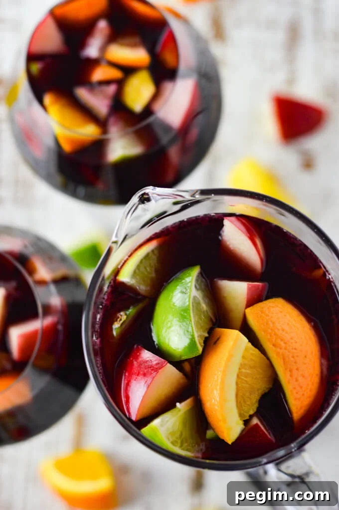 A photo from above of a pitcher of red wine sangria with chunks of fresh apples, oranges, lemons and limes. Two glasses filled with the sangria sit beside the pitcher. Chucks of the fresh fruit are scattered on the board below.
