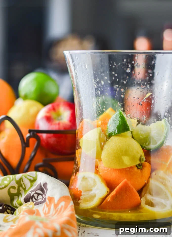 A pitcher of fresh squeezed oranges, lemons and limes with a colorful napkin beside it. A metal basket with fresh fruit sits in the background.