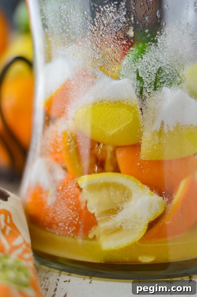 A pitcher of fresh squeezed oranges, lemons and limes with a colorful napkin beside it. A metal basket with fresh fruit sits in the background.
