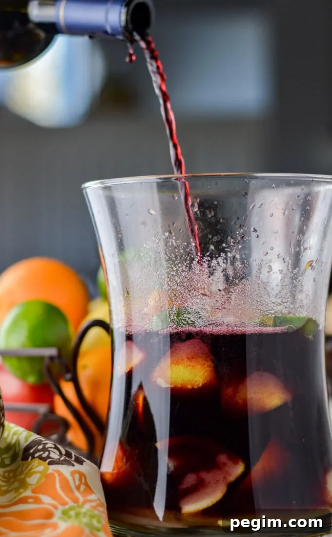 A bottle of red wine is being poured into a pitcher of chunks of fresh fruits. A basket of whole fresh apples, oranges, limes and lemons sit in the background.