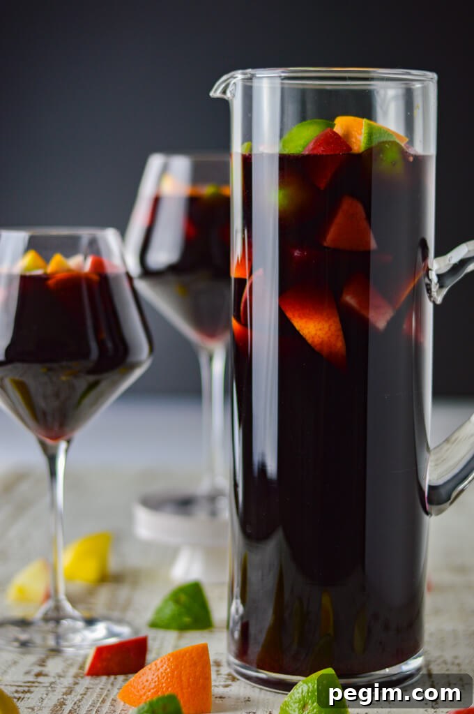 A pitcher of red wine sangria with chunks of fresh apples, oranges, lemons and limes. Two glasses filled with the sangria sit beside the pitcher. Chucks of the fresh fruit are scattered on the board below.