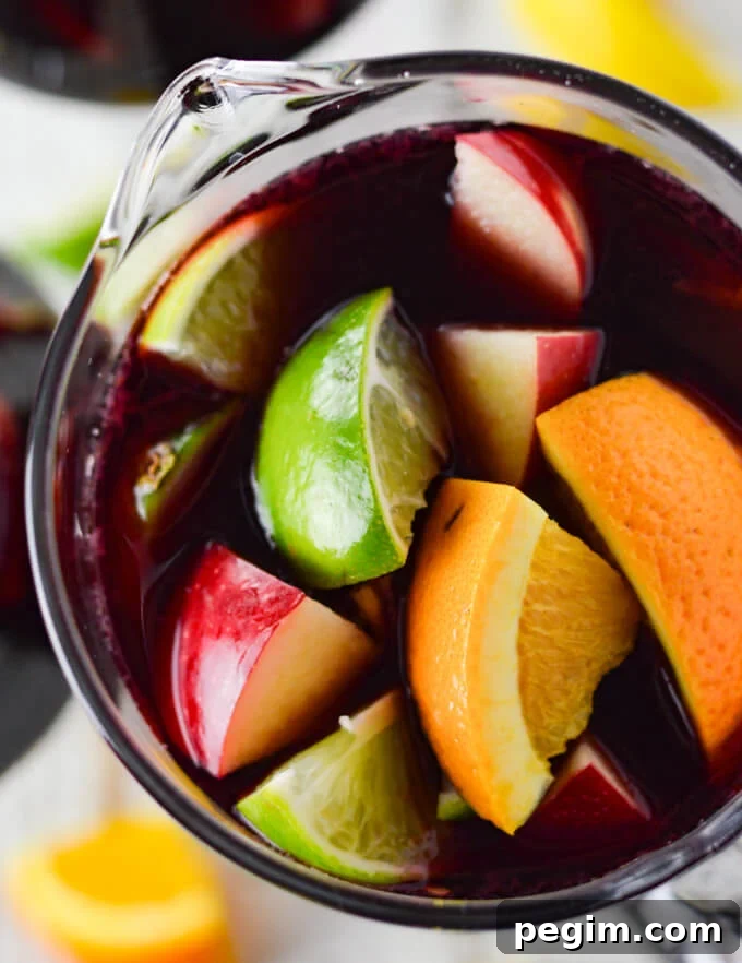 A closeup photo from above of a pitcher of red wine sangria with chunks of fresh apples, oranges, lemons and limes. Two glasses filled with the sangria sit beside the pitcher. Chucks of the fresh fruit are scattered on the board below.