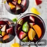 A photo of a pitcher of red wine sangria with chunks of fresh apples, oranges, lemons and limes. Two glasses filled with the sangria sit beside the pitcher. Chucks of the fresh fruit are scattered on the board below.