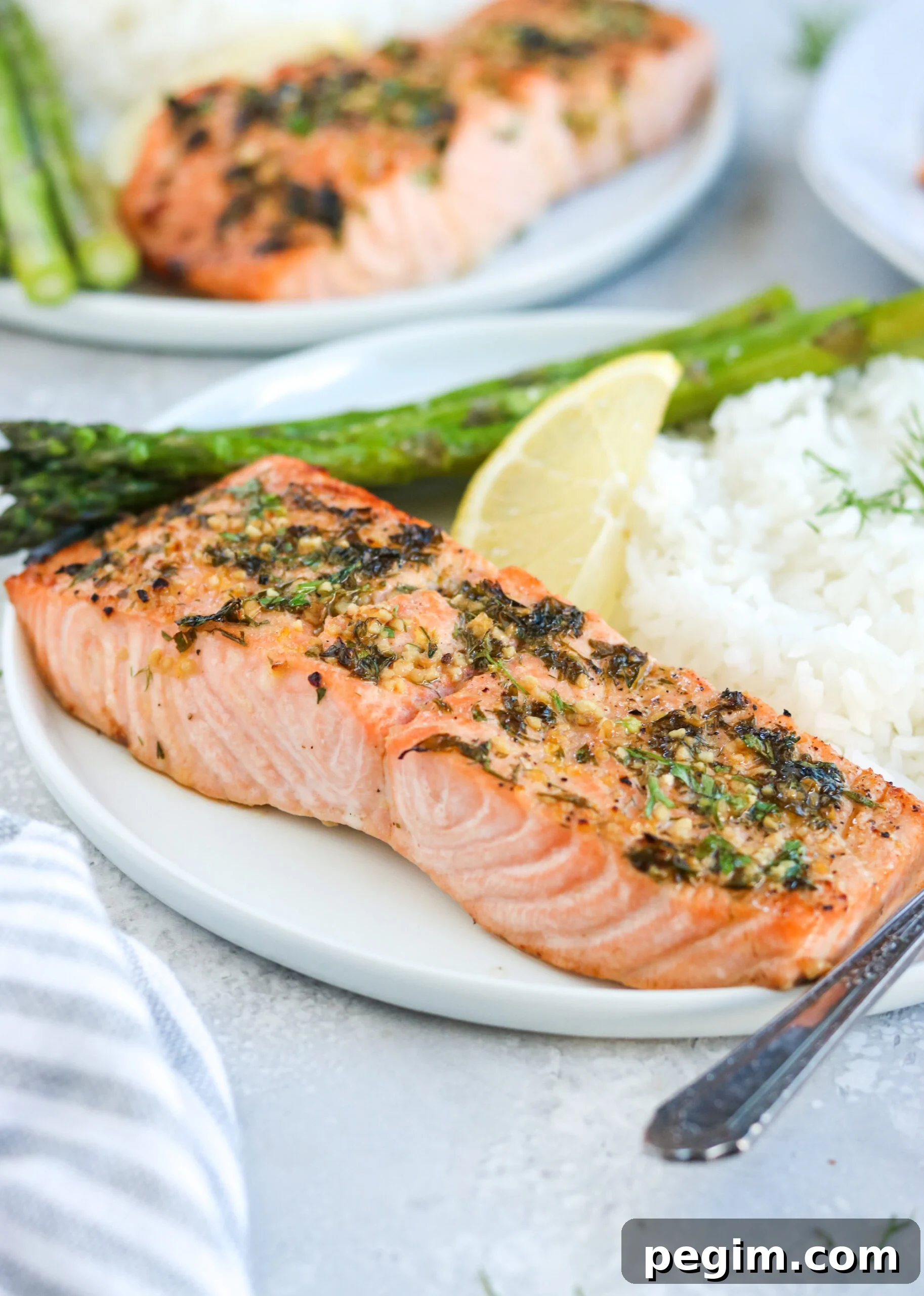 side view of garlic herb baked salmon on a white plate with asparagus, white rice, and a lemon wedge, next to a silver fork with a white background side view of garlic herb baked salmon on a white plate with asparagus, white rice, and a lemon wedge, next to a silver fork with a white background