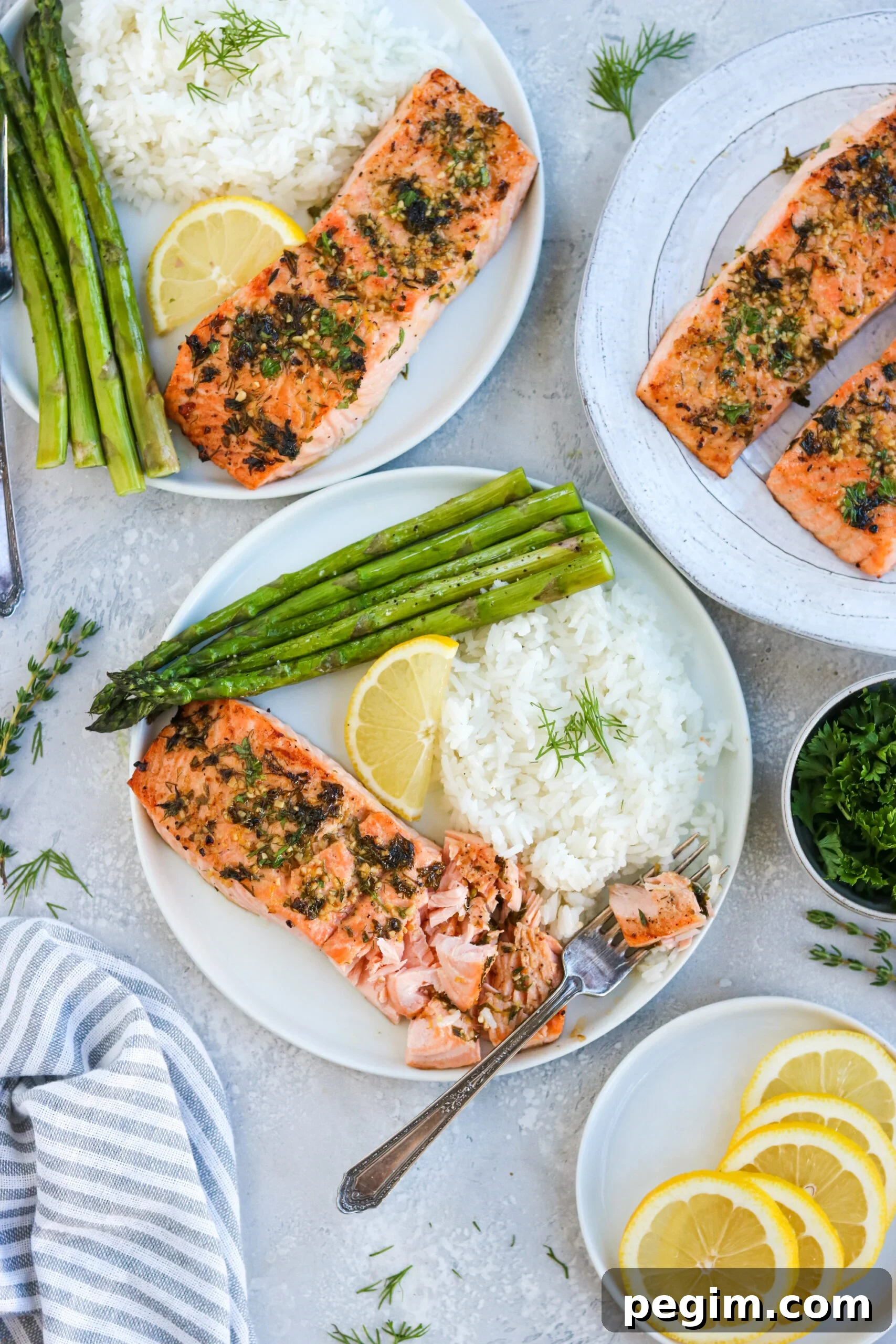 three plates of garlic herb baked salmon on a white plate with asparagus, white rice, and a lemon wedge, next to a silver fork with a white background three plates of garlic herb baked salmon on a white plate with asparagus, white rice, and a lemon wedge, next to a silver fork with a white background