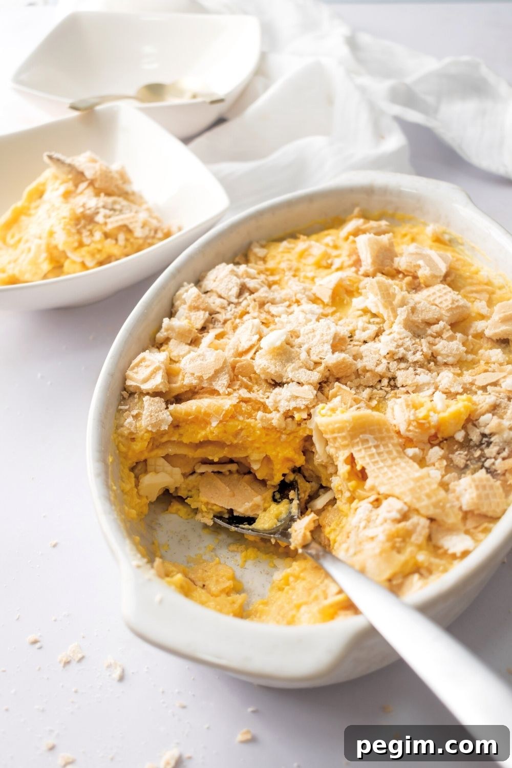 A white casserole dish that is filled with banana pudding. There is a spoon in the front of the dish with a scoop of banana pudding out of i. The scoop is behind the casserole dish in a square bowl.