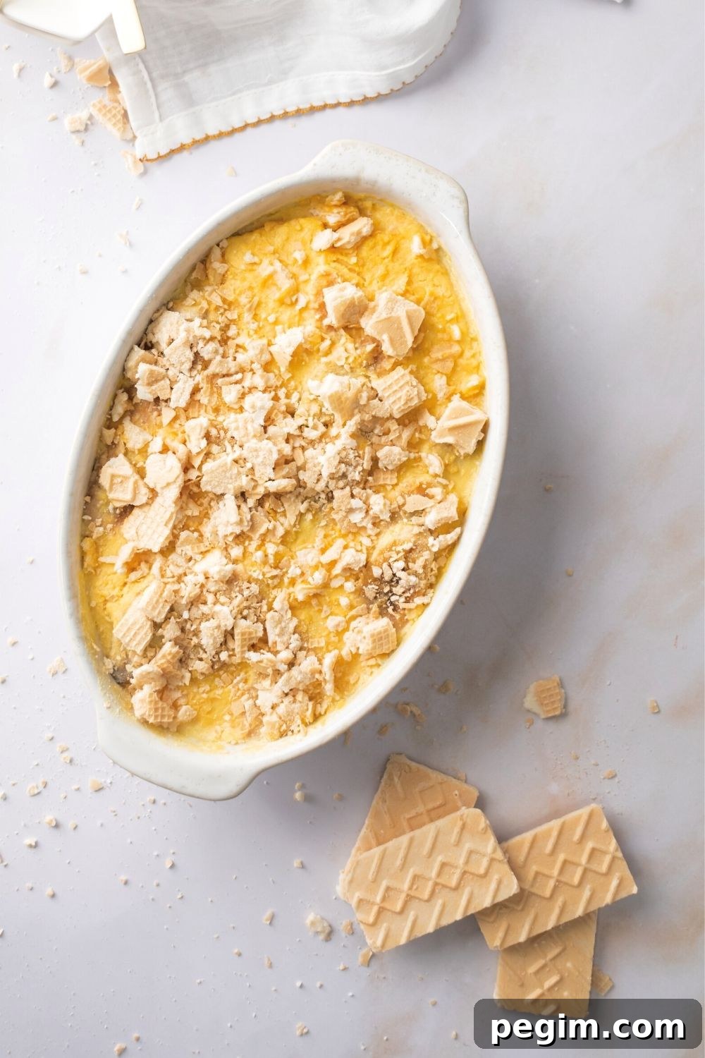 Crushed up vanilla wafer cookies on top of banana pudding in a white casserole dish. In front of it is a few squares of vanilla wafer cookies.