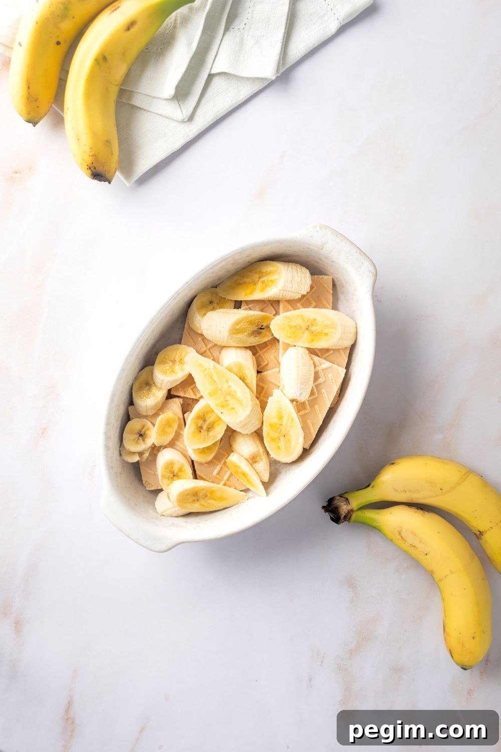 Banana slices on top of vanilla wafer cookies in a white casserole dish. There are two bananas in front of the dish and part of two bananas behind the dish.