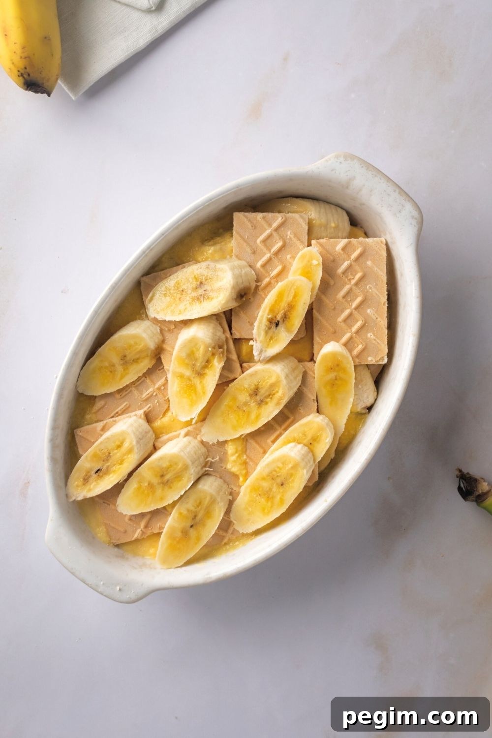 A couple of banana slices on top of vanilla wafer cookies in a white casserole dish on a white counter.