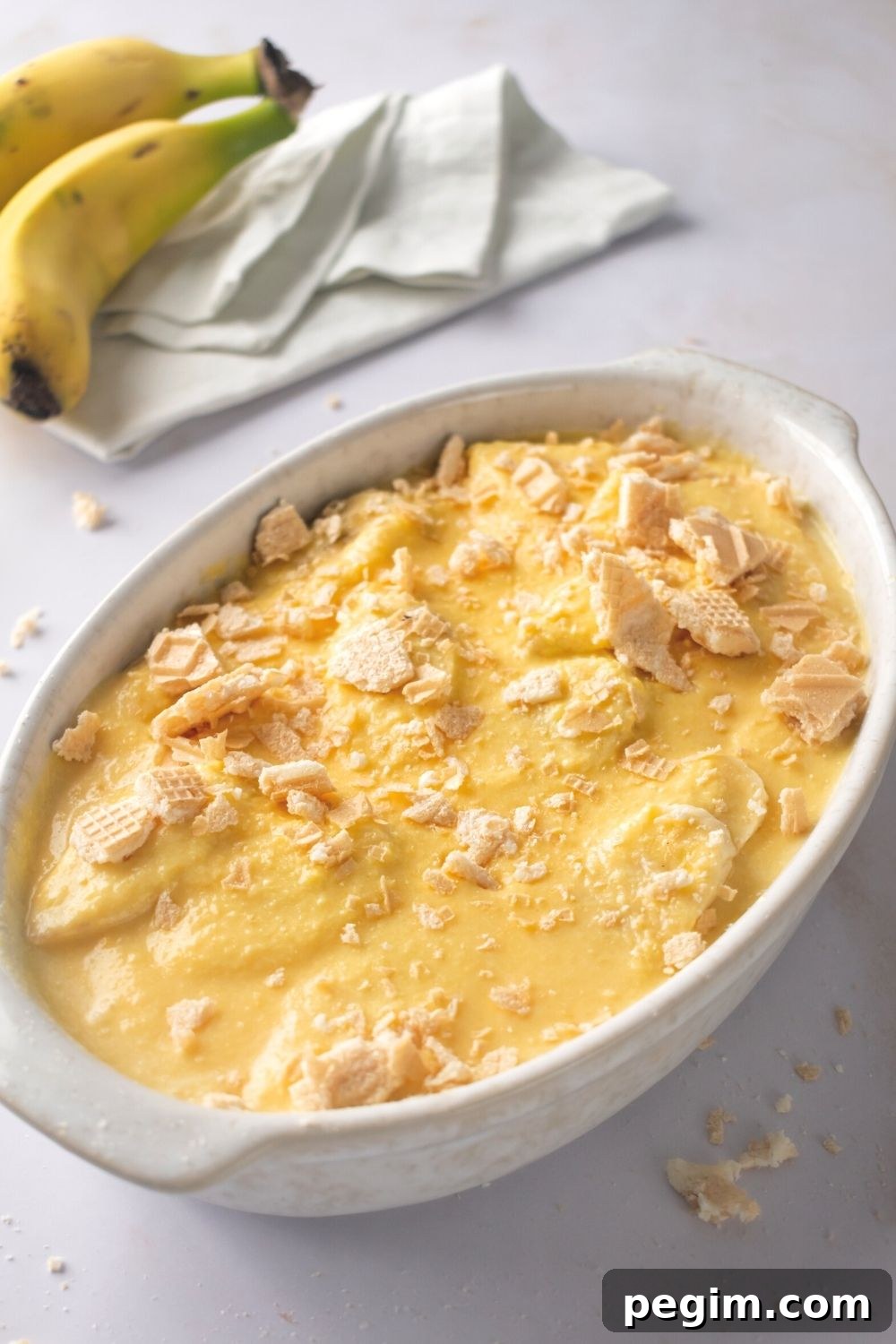 A white casserole dish filled with banana pudding. Behind it is part of two bananas.