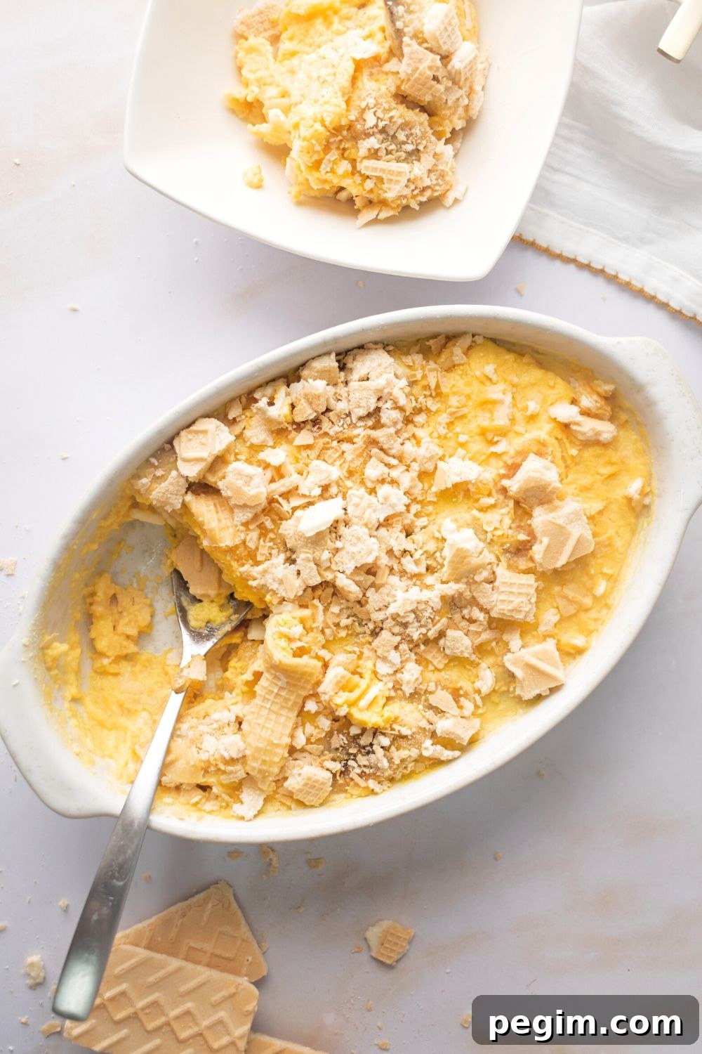 Crushed up vanilla wafer cookies on top of banana pudding in a white casserole dish. A spoon is in the dish to the left of the pudding with a scoop taken out of that section. Behind it is the scoop of pudding in part of a square bowl.
